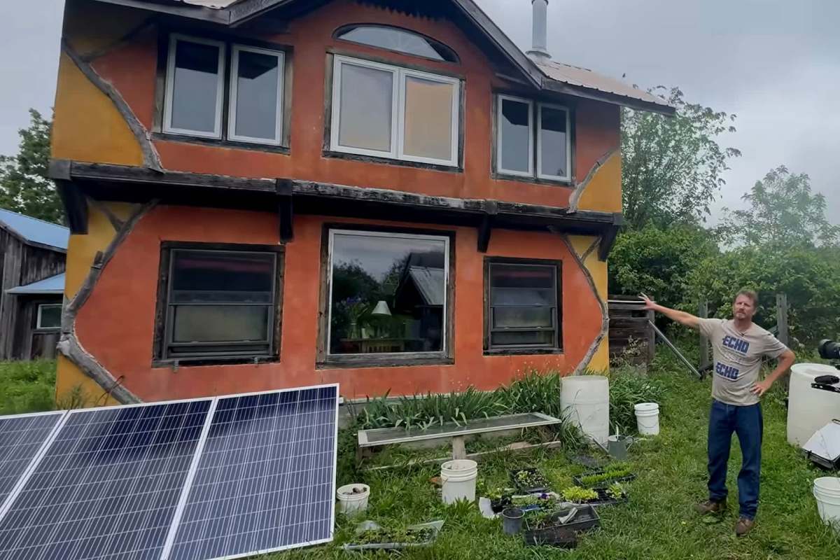 A man stands next to a colorful house with solar panels and plants in the foreground.