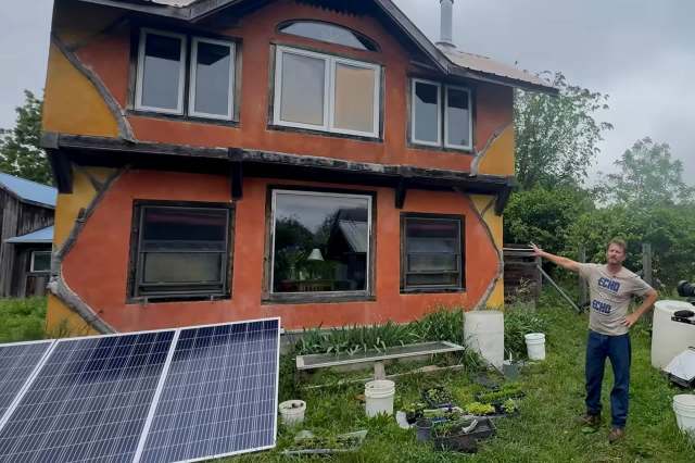 A man stands next to a colorful house with solar panels and plants in the foreground.