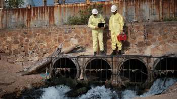 Two workers in yellow protective suits stand by a water outlet, examining a device and a toolbox.