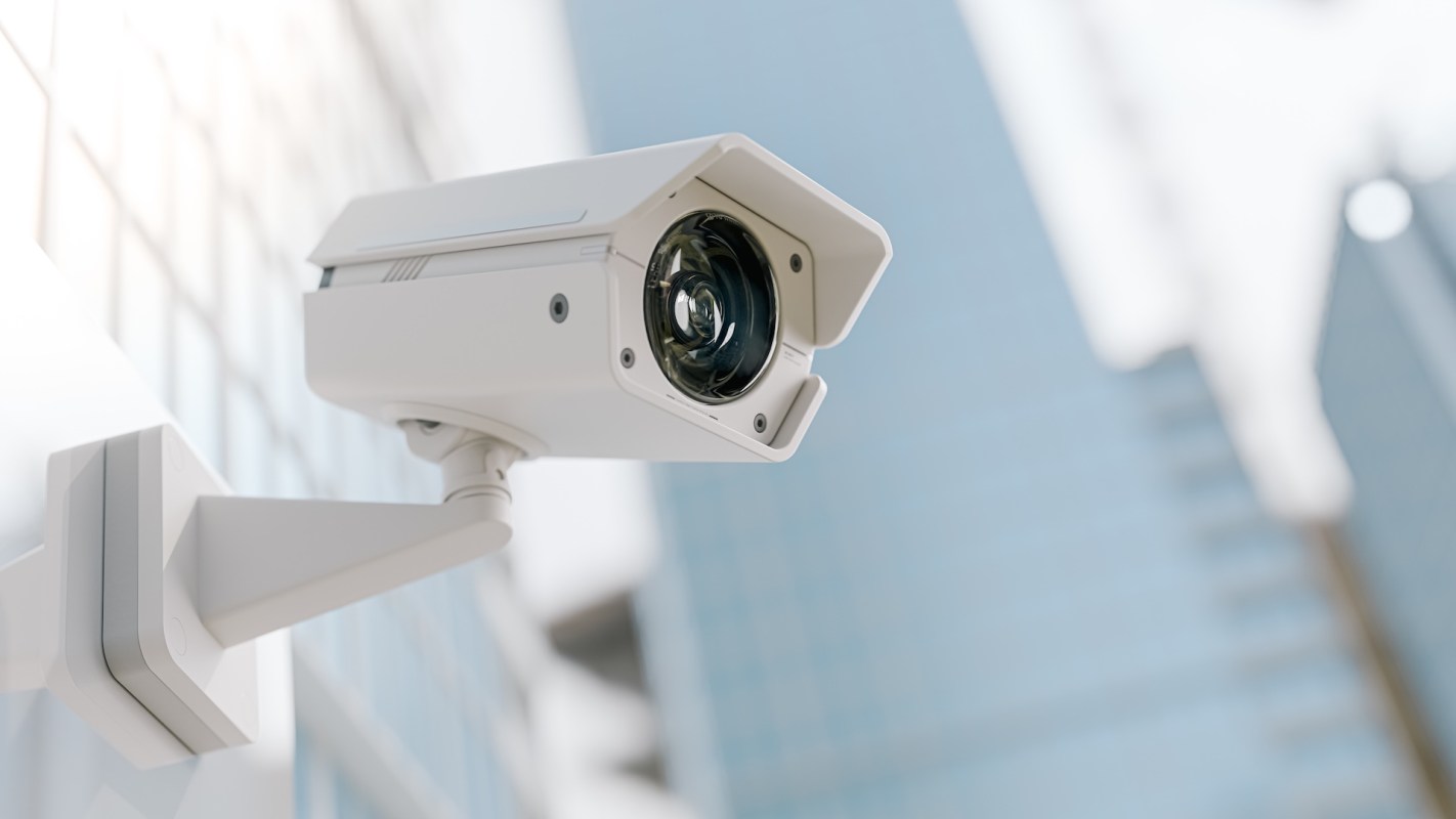 A security camera mounted on a wall with a blurred building in the background.
