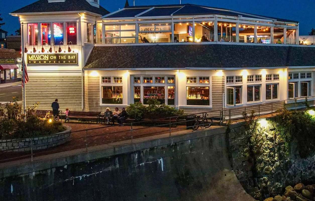 A restaurant building with a balcony, outdoor seating, and illuminated signage by the water at night.