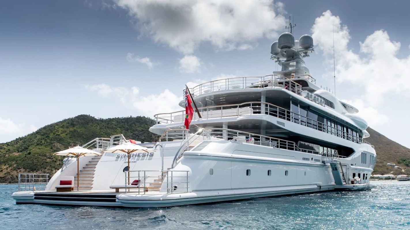 A large luxury yacht anchored near a hilly coastline with blue water and a cloudy sky.
