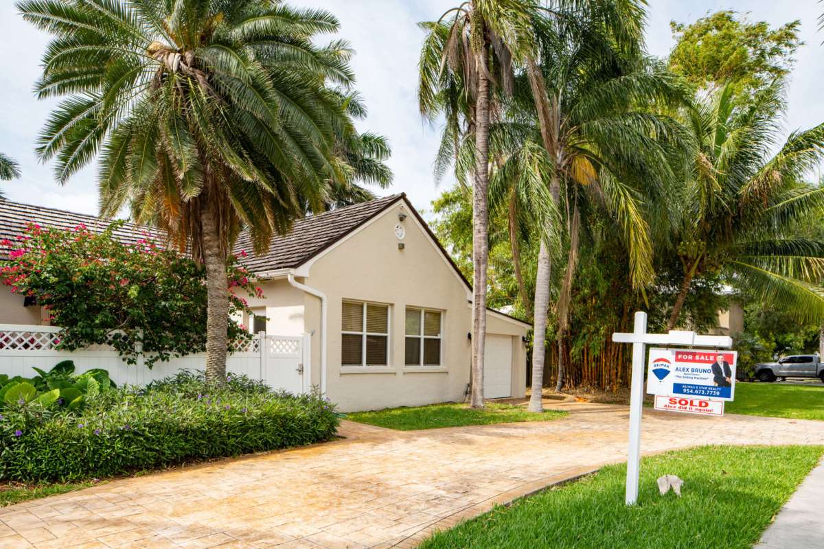 A house with a "For Sale" sign surrounded by palm trees and a landscaped yard.