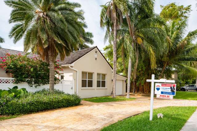 A house with a "For Sale" sign surrounded by palm trees and a landscaped yard.
