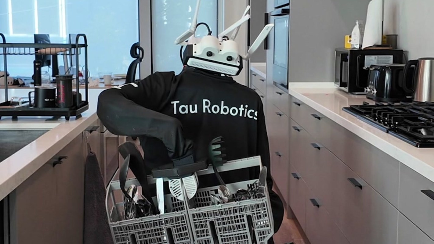 A robot wearing a "Tau Robotics" shirt holds a basket of kitchen utensils in a modern kitchen.