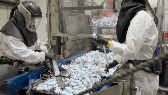 Two workers in protective suits handle canisters from an EV battery on a conveyor belt in a processing facility.