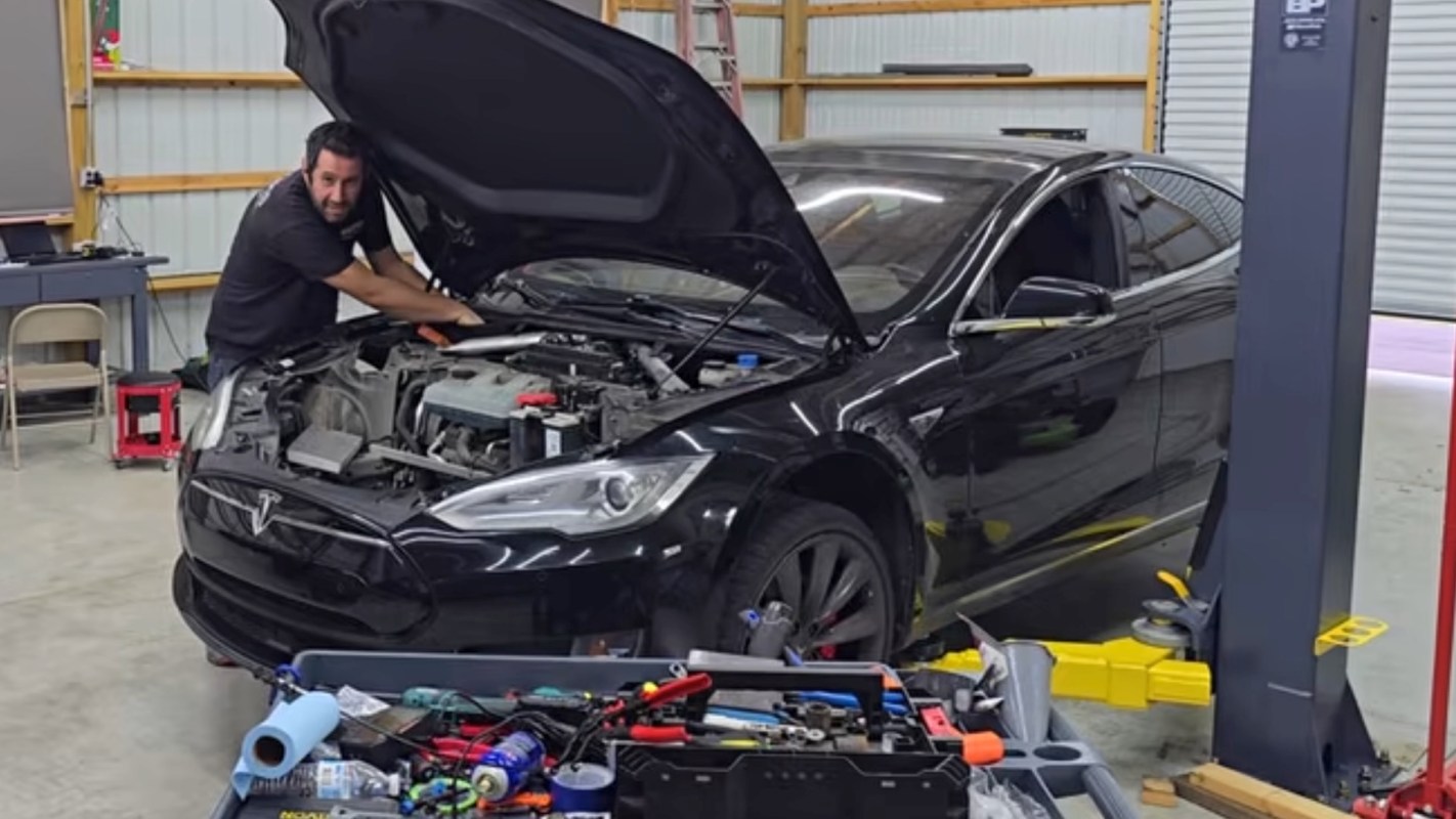 A person works on a Tesla car's engine in a garage filled with tools and equipment.