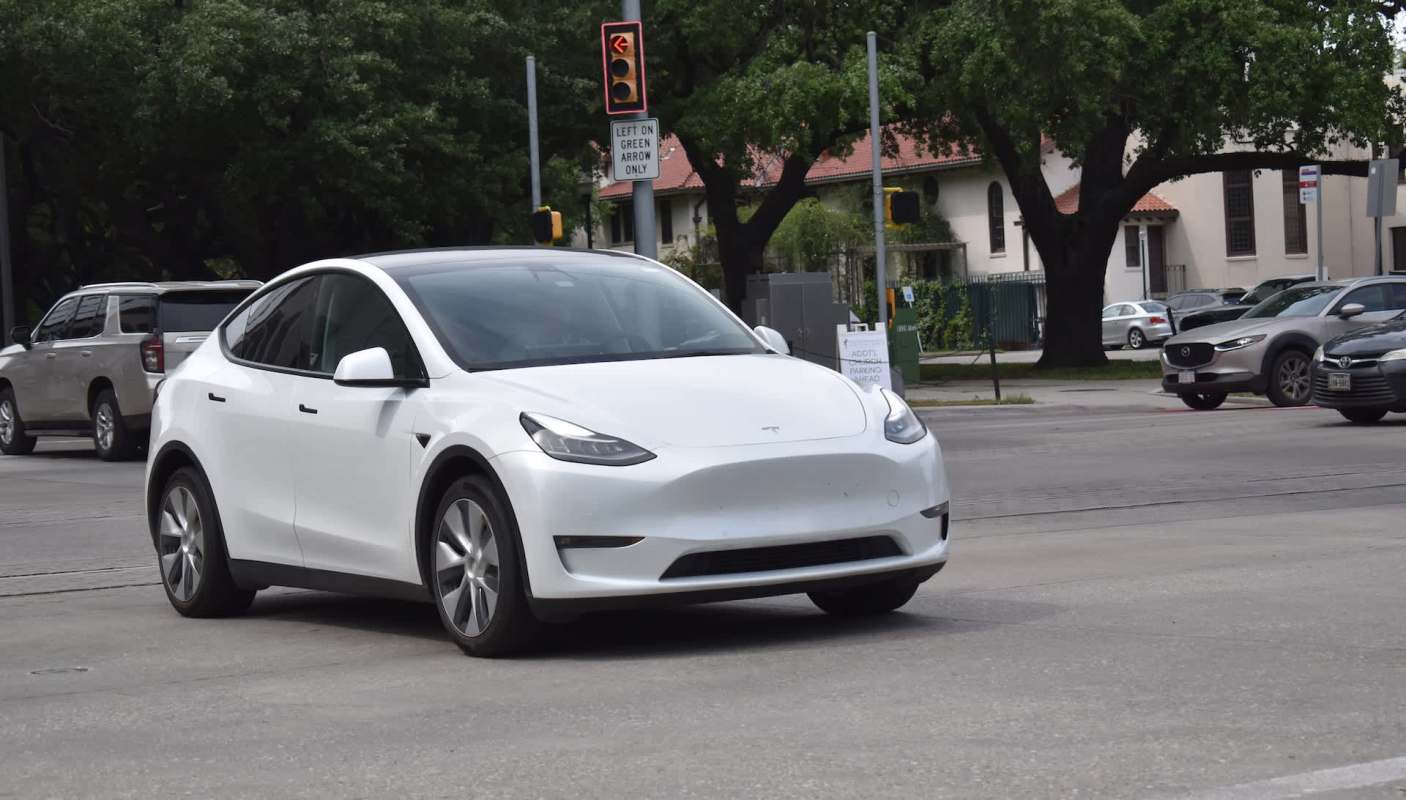 A white Tesla Model Y drives through an intersection with traffic signals and nearby cars.