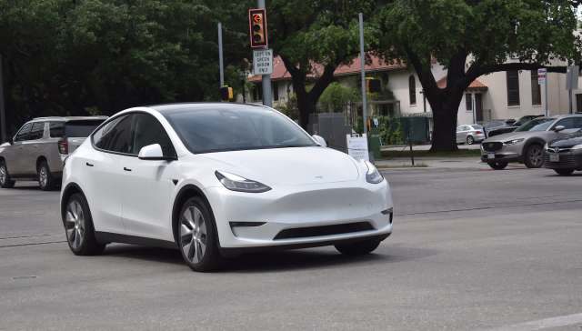 A white Tesla Model Y drives through an intersection with traffic signals and nearby cars.