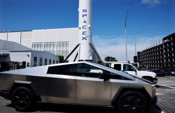 A futuristic vehicle parked near a SpaceX rocket in an industrial area.