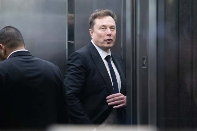 Elon Musk in a suit stands in an elevator next to another man.