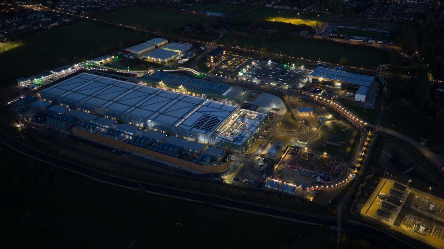 An aerial view of an industrial complex at night, illuminated with various lights and surrounded by green fields.