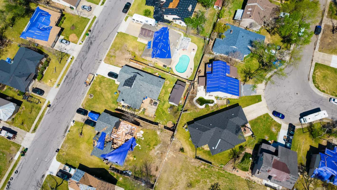 An aerial view of a neighborhood with several houses, some under construction and covered with blue tarps after tornado damage.