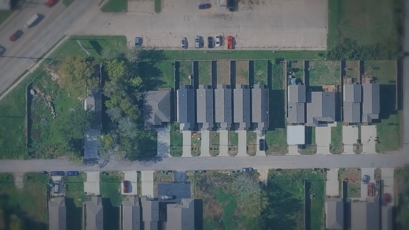 An overhead view of a residential area with houses, parking spaces, and tree-lined streets.
