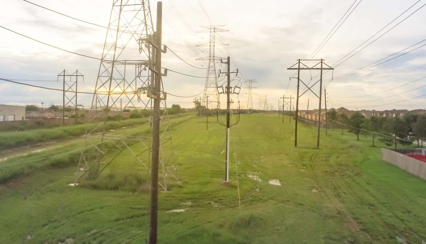 Power lines stretch across a grassy area with homes visible in the background under a cloudy sky.