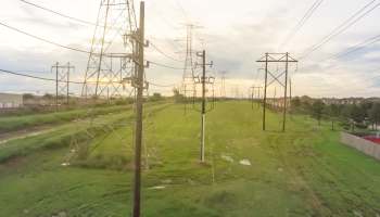Power lines stretch across a grassy area with homes visible in the background under a cloudy sky.