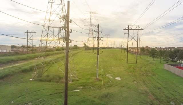 Power lines stretch across a grassy area with homes visible in the background under a cloudy sky.