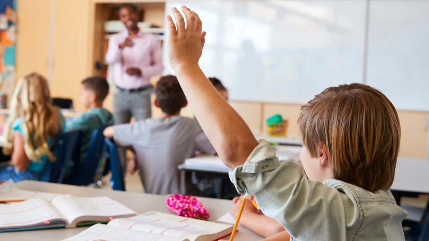 A student raises their hand in a classroom while a teacher speaks to the class.