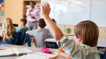 A student raises their hand in a classroom while a teacher speaks to the class.