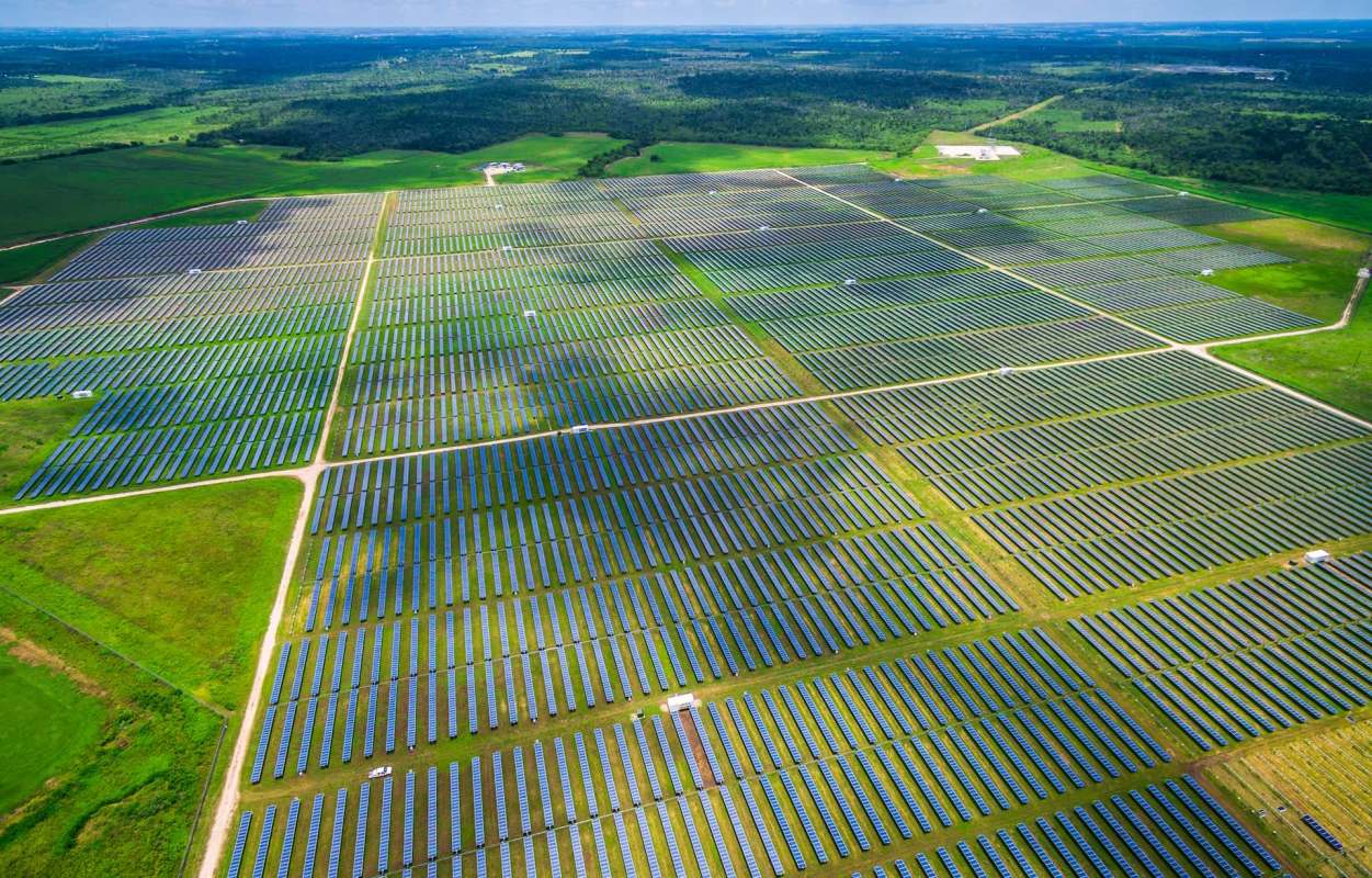 Aerial view of a large solar farm with numerous solar panels arranged in rows across a green landscape.