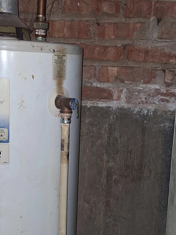 A water heater against a brick wall with a visible valve and maintenance instructions on the side.