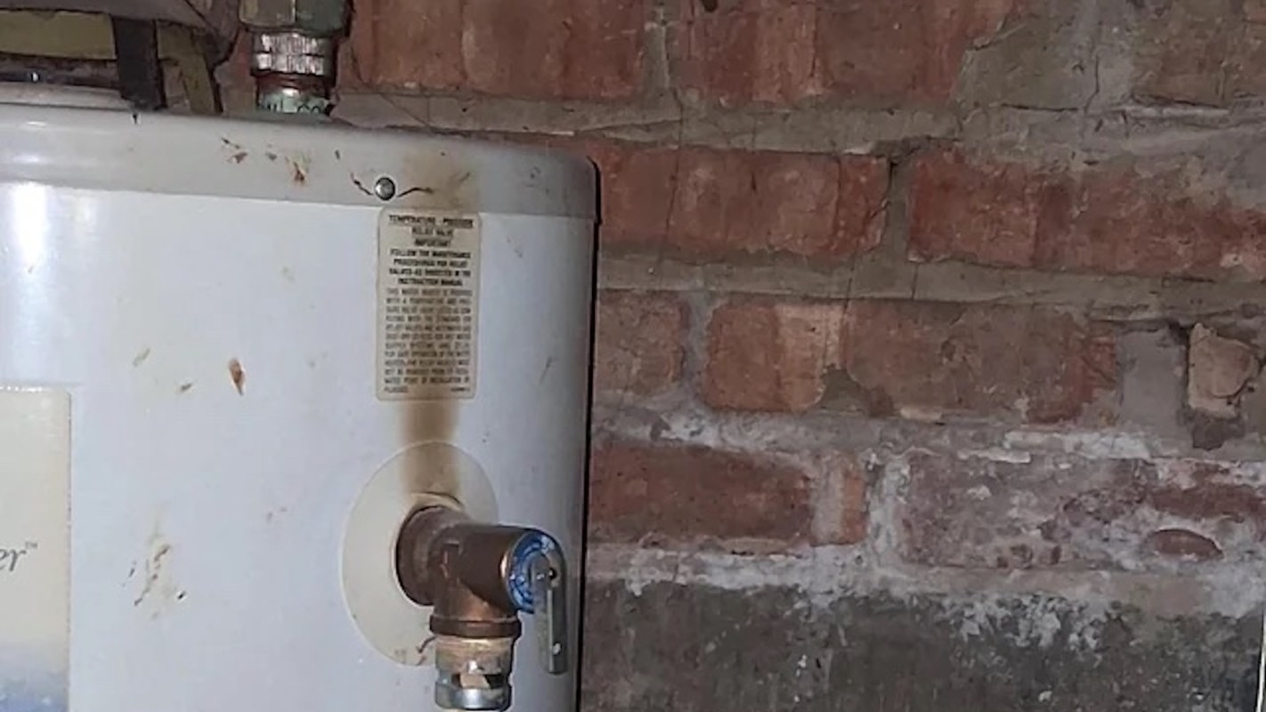 A water heater against a brick wall with a visible valve and maintenance instructions on the side.