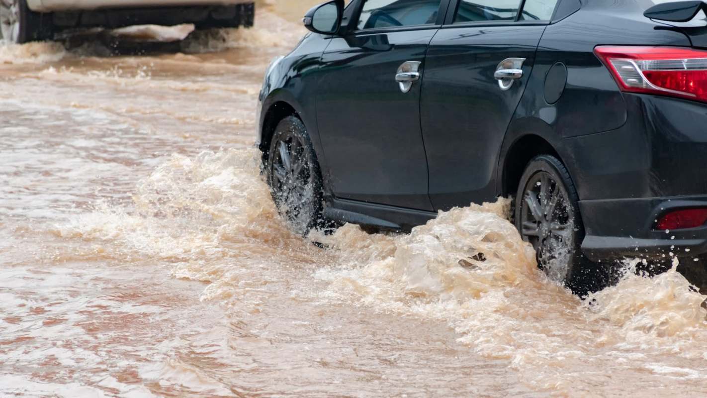 A black car driving through a flooded street, with water splashing around its wheels.