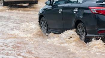 A black car driving through a flooded street, with water splashing around its wheels.