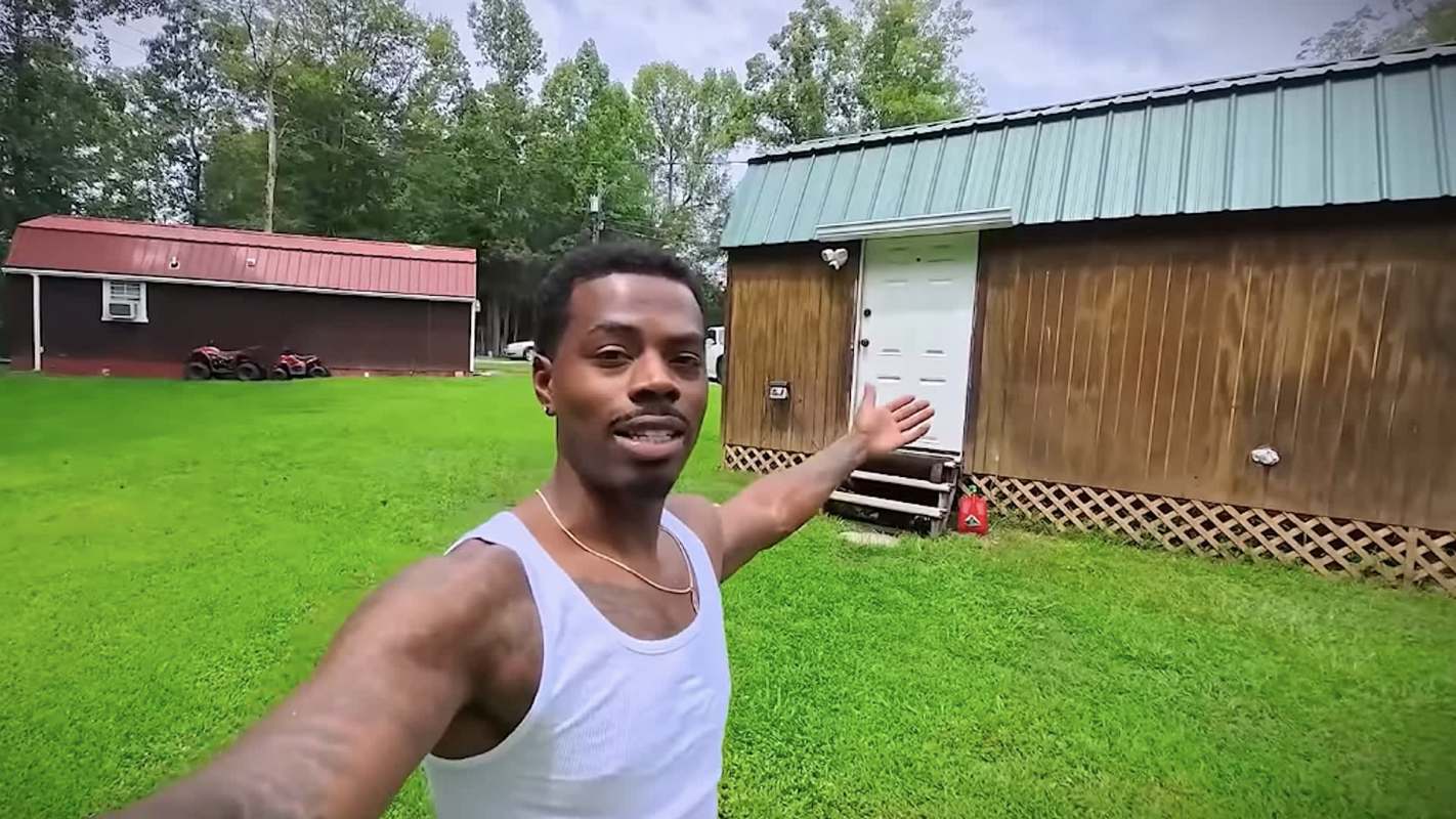 A man stands in a green yard, gesturing towards a wooden building with a metal roof.