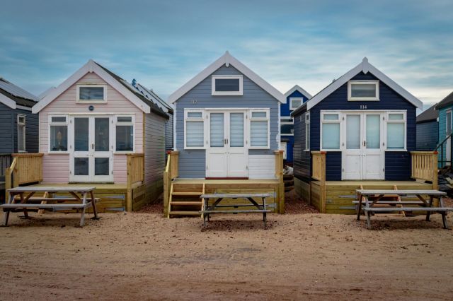 Three tiny homes in pastel colors with wooden steps and picnic tables in front, set against a sandy ground.