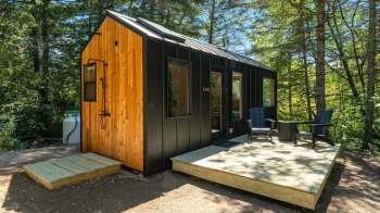 A modern black and wood cabin surrounded by trees, featuring a small deck and outdoor seating.