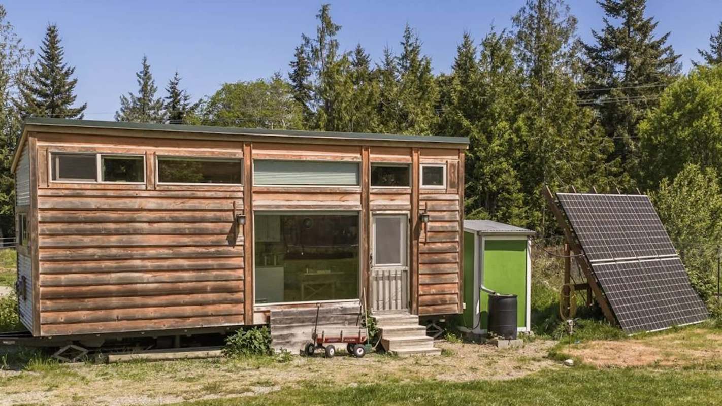 A tiny house with a wood exterior and solar panels in a grassy area surrounded by trees.