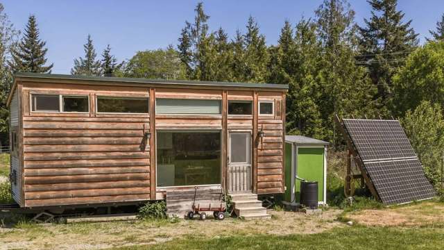 A tiny house with a wood exterior and solar panels in a grassy area surrounded by trees.