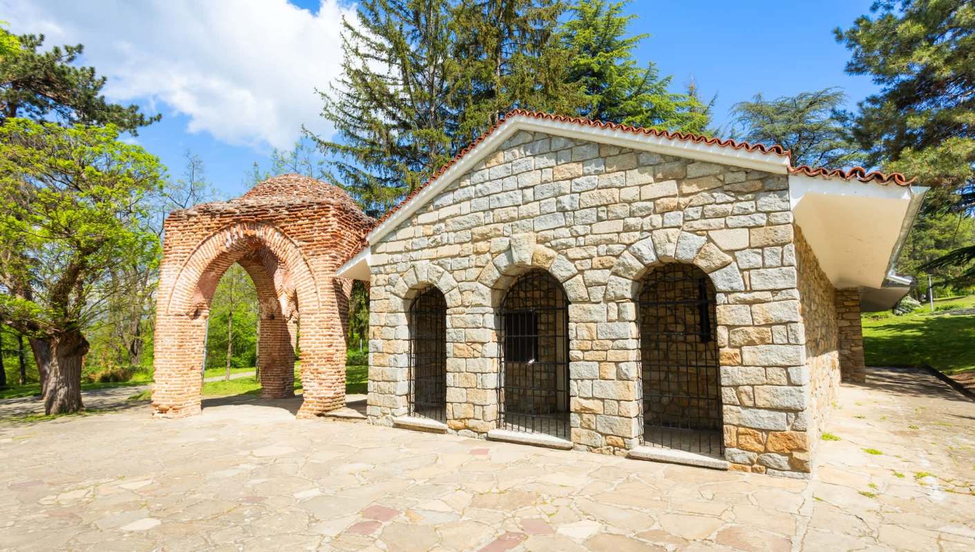 A stone building with arched windows and a brick structure beside it, surrounded by trees.