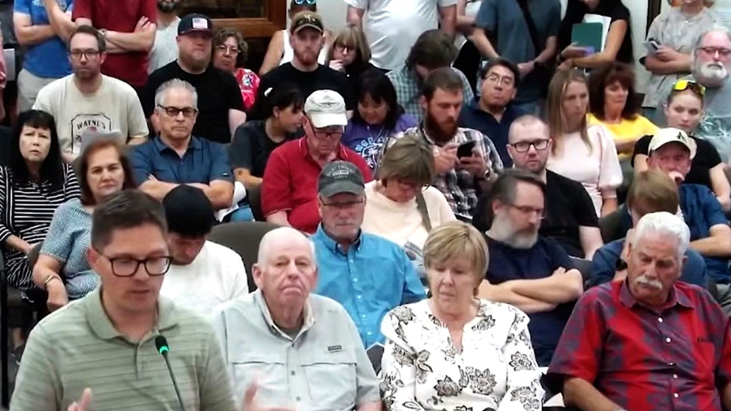 A group of people attending a public meeting, some seated and others standing, focused on a speaker at the front.