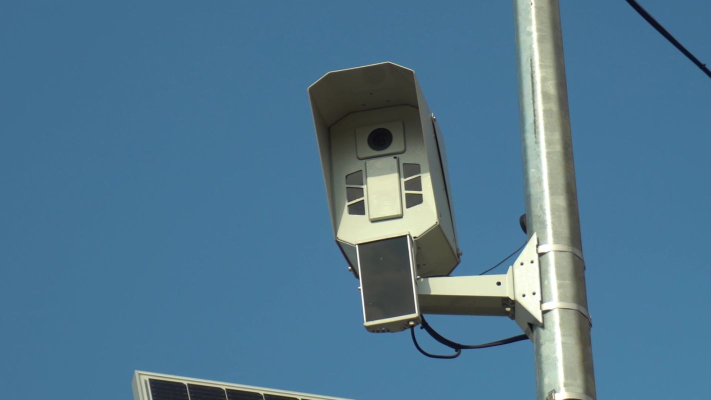 A mounted surveillance camera attached to a metal pole against a clear blue sky.