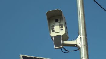 A mounted surveillance camera attached to a metal pole against a clear blue sky.