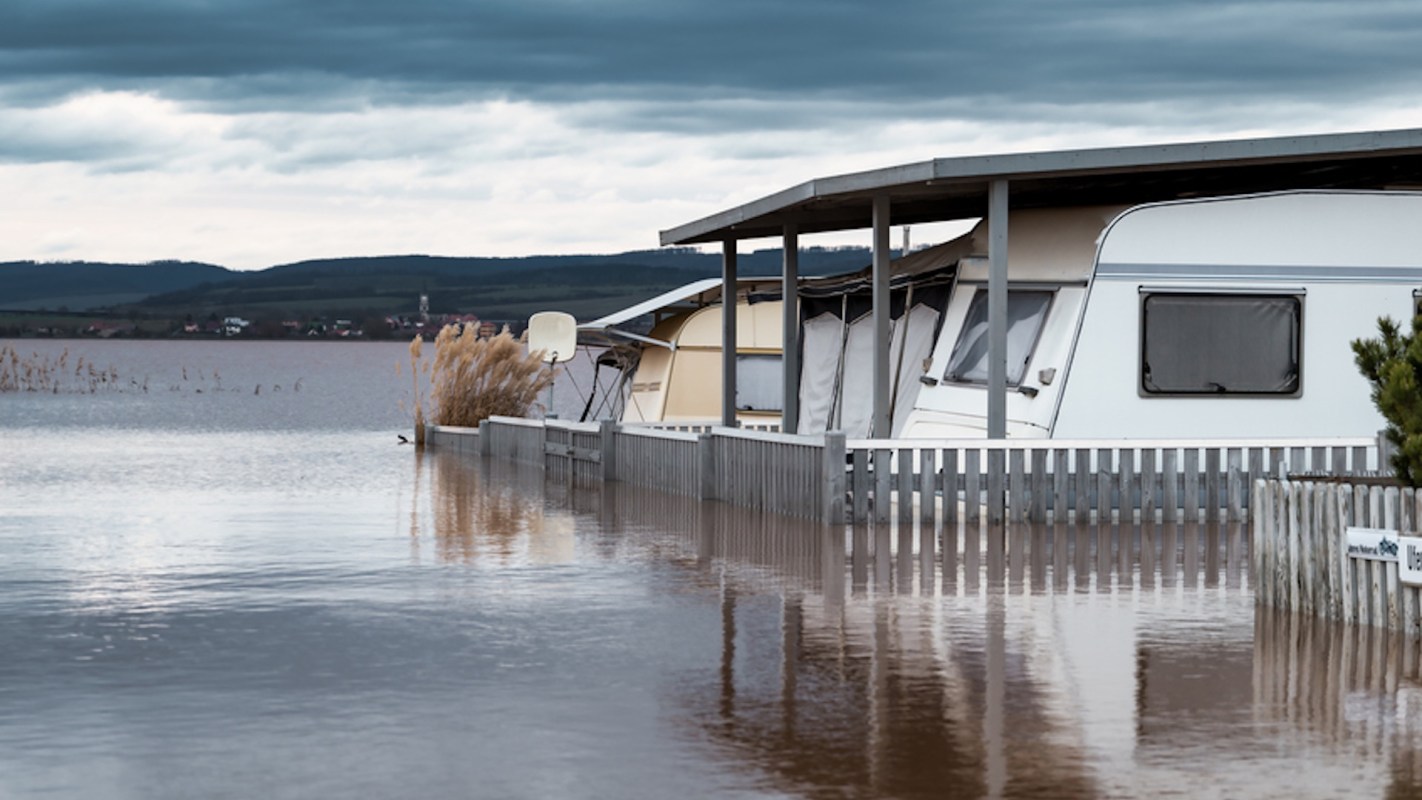A partially submerged caravan next to a flooded area with tall grasses and cloudy skies.