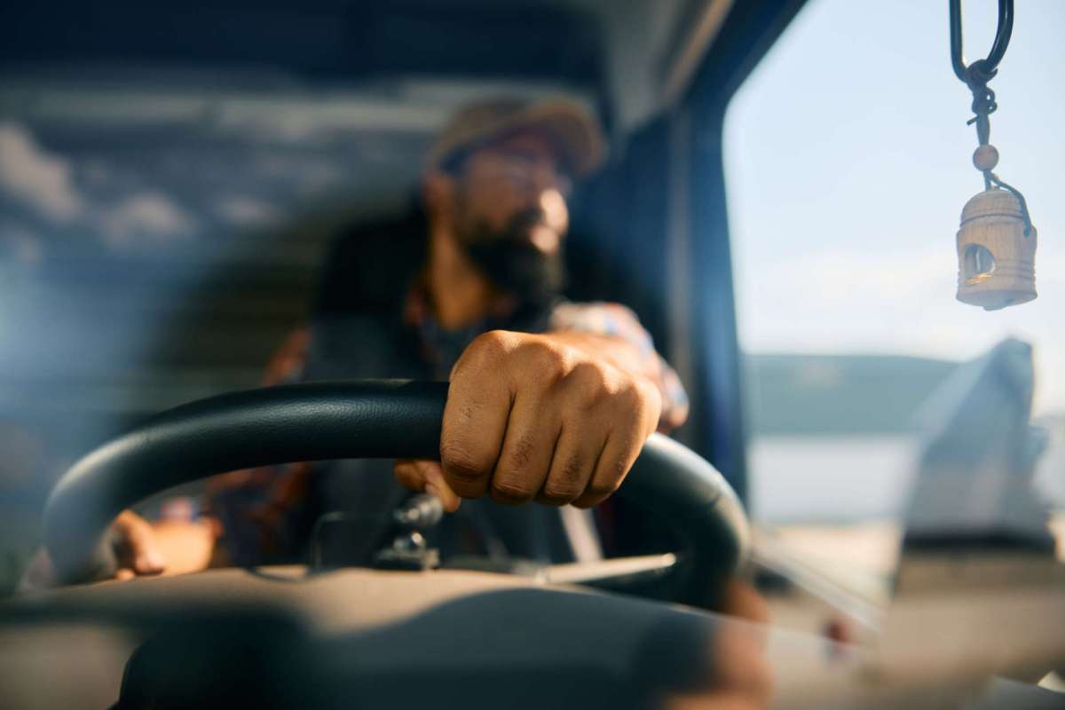 A person drives a vehicle, with their hand on the steering wheel and a decorative item hanging in the background.