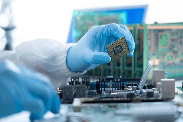 A person in blue gloves holds a computer chip above a circuit board.