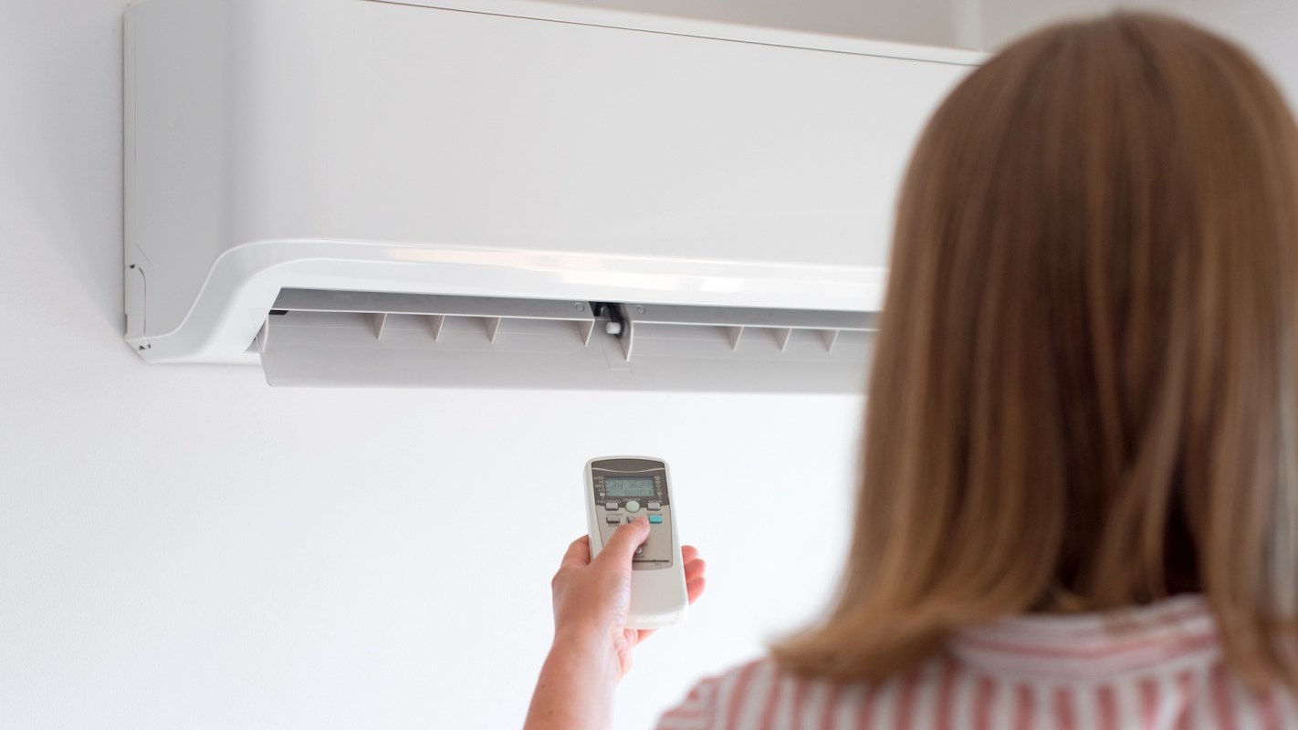 A person holds a remote control aimed at a wall-mounted air conditioning unit.