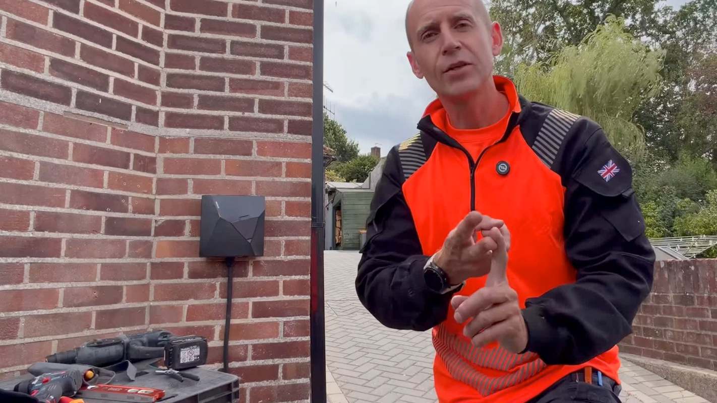 A man in an orange jacket gestures while standing by a brick wall and tools on a work surface.