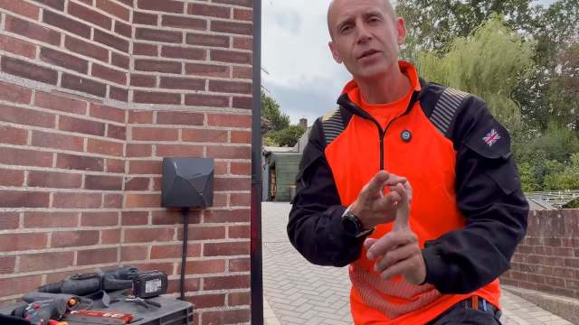 A man in an orange jacket gestures while standing by a brick wall and tools on a work surface.