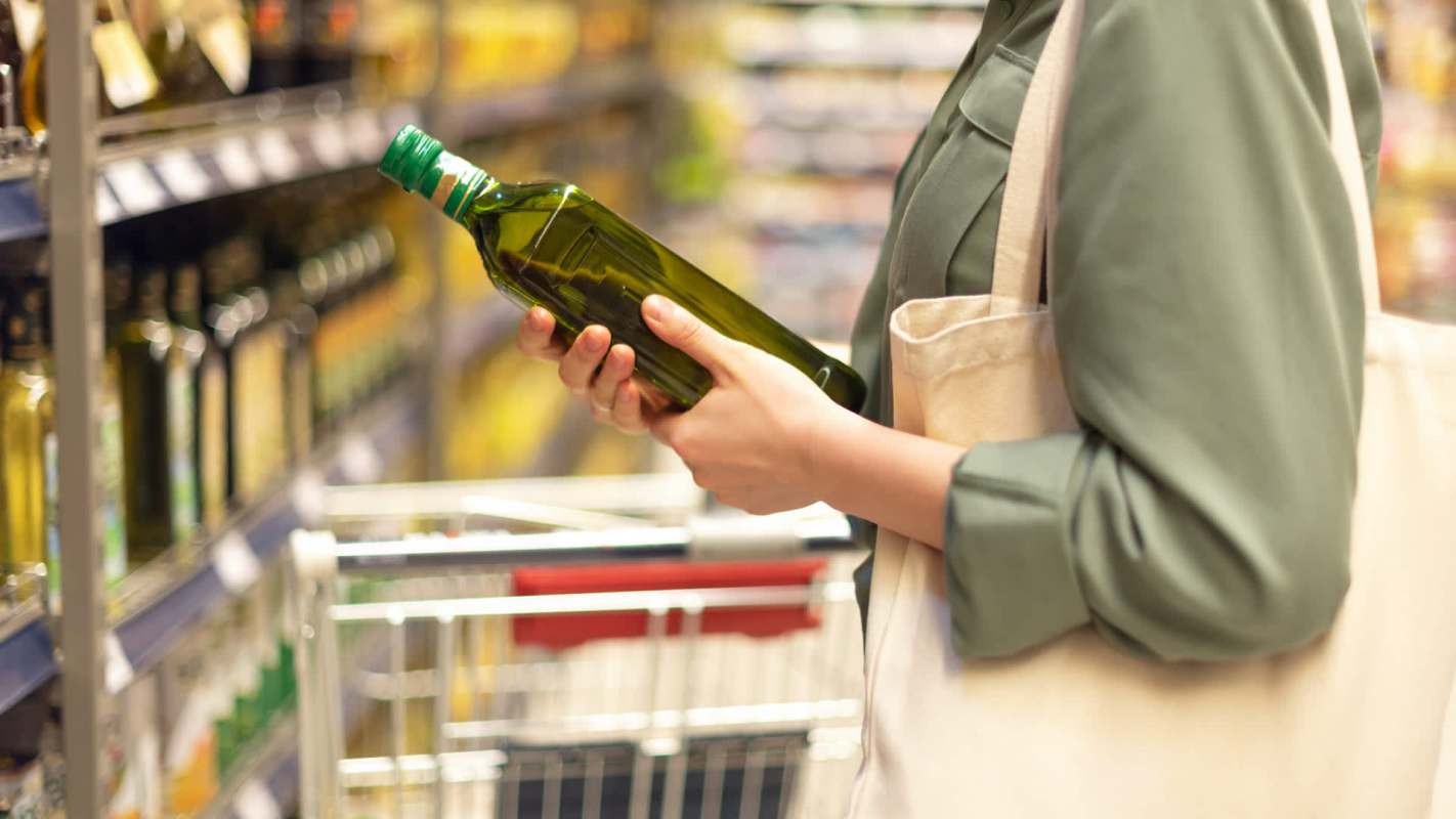 A person holds a bottle of olive oil while shopping in a grocery store aisle.