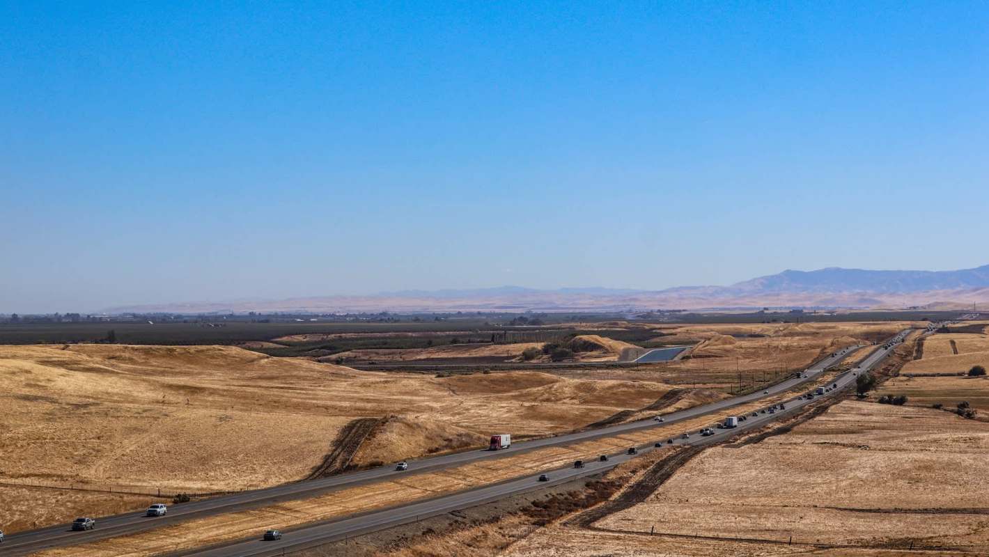 A highway runs through dry, grassy hills with distant mountains under a clear blue sky.