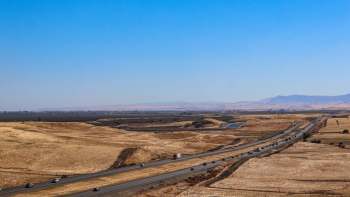 A highway runs through dry, grassy hills with distant mountains under a clear blue sky.