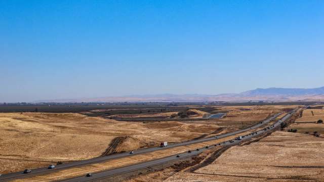 A highway runs through dry, grassy hills with distant mountains under a clear blue sky.