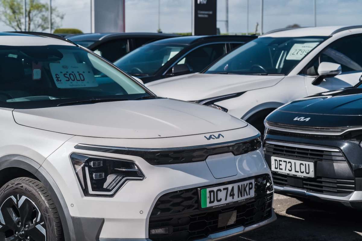 A white Kia car in a parking lot with other vehicles in the background, one marked as sold.