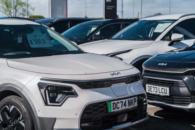 A white Kia car in a parking lot with other vehicles in the background, one marked as sold.