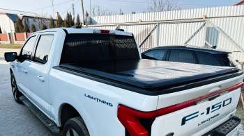 A white Ford F-150 Lightning truck parked near a building with a corrugated metal fence and another vehicle in the background.
